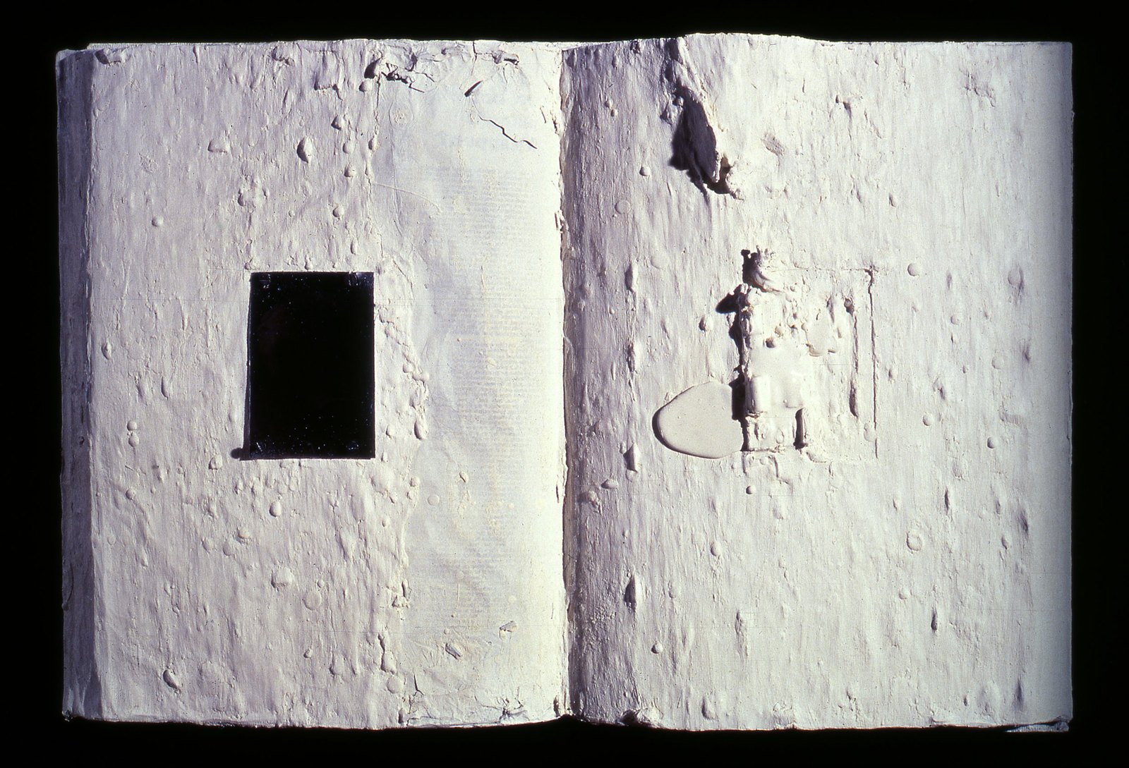 A white encrusted book-object, open, with a black rectangle set into one page.