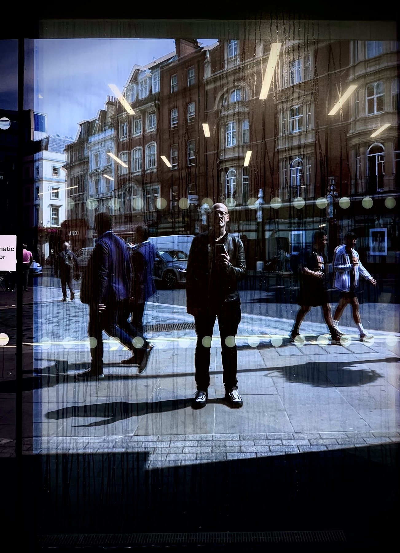 No End In Sight 025: The artist with a phone reflected in a dotted-glass window with walking pedestrians on either side and a sunlit London street.
