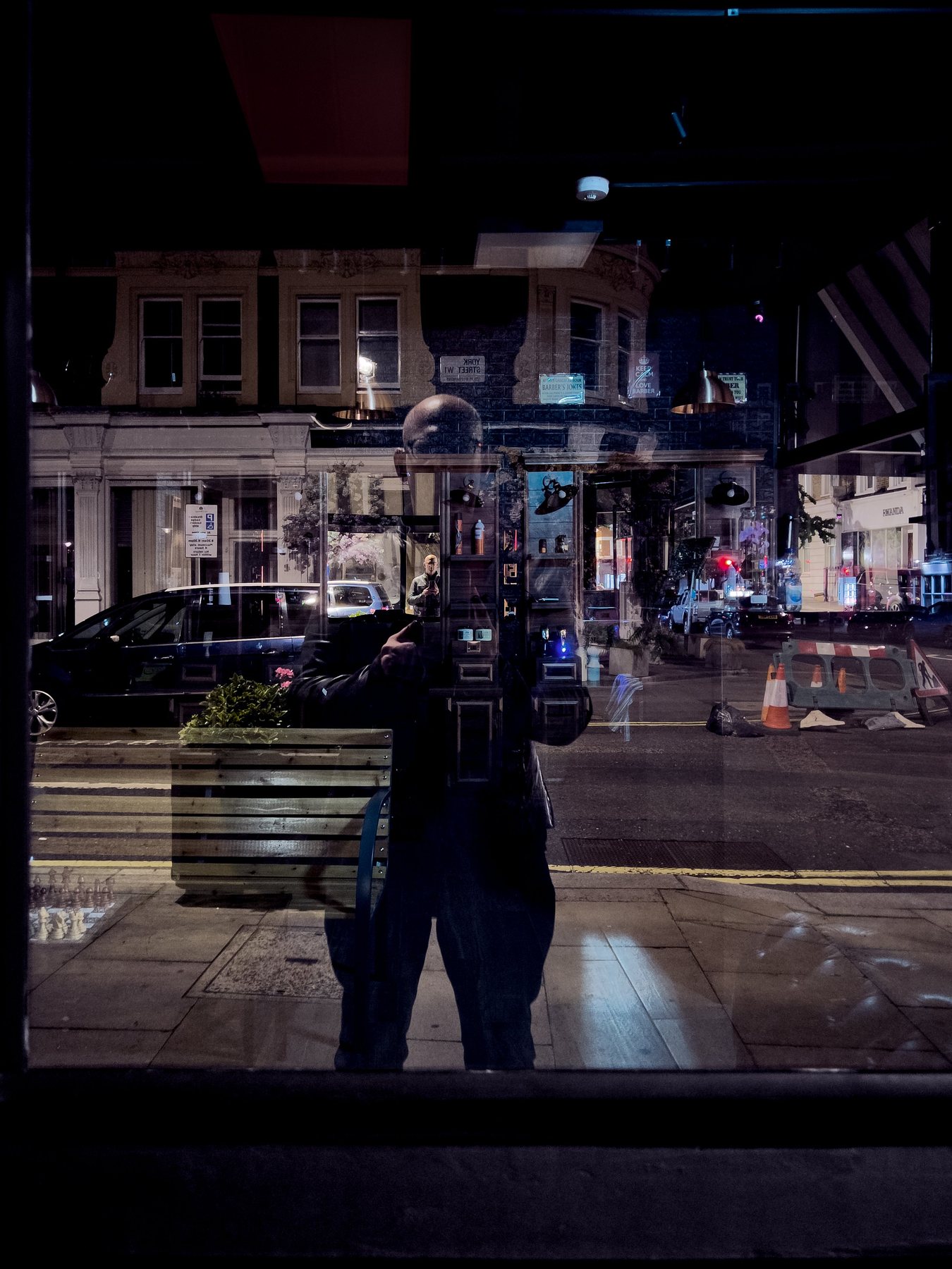 No End In Sight 035: A night window reflecting a lit London street, orange traffic cones and parked cars, with the artist's dark figure and a planter.
