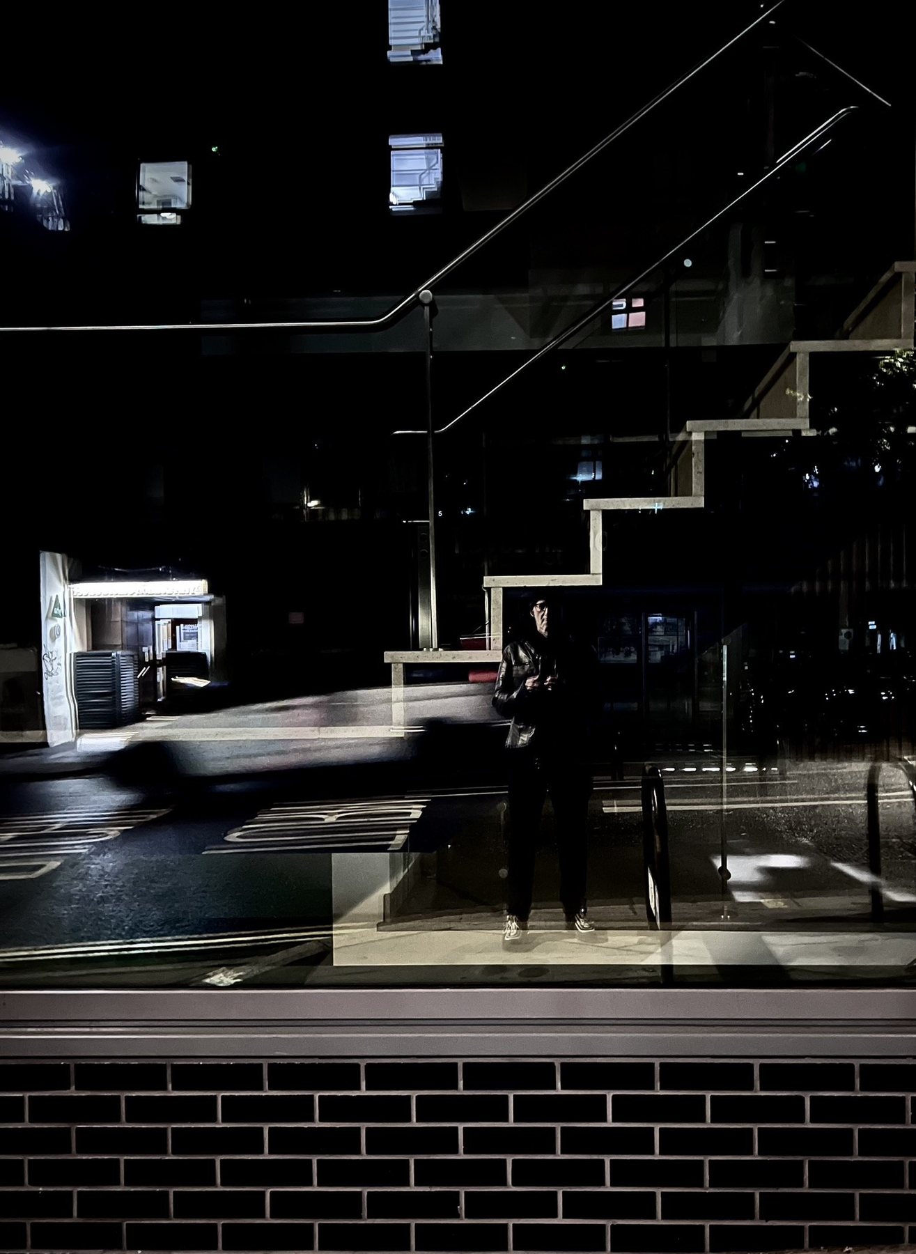No End In Sight 018: A dark night window reflecting an angular staircase, with the artist and a blurred passing car, above a black brick base.