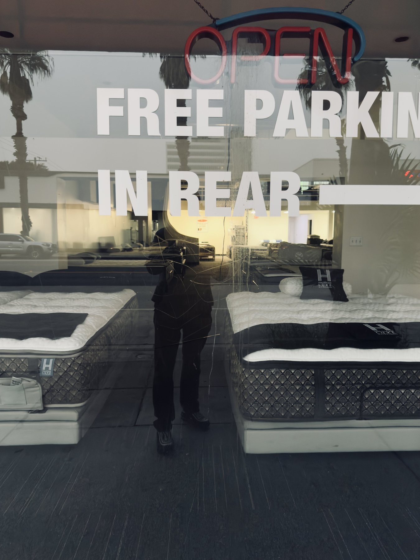 No End In Sight 126: The artist's dark reflection in a shop window under a glowing OPEN sign and the words FREE PARKING IN REAR, with mattresses inside.
