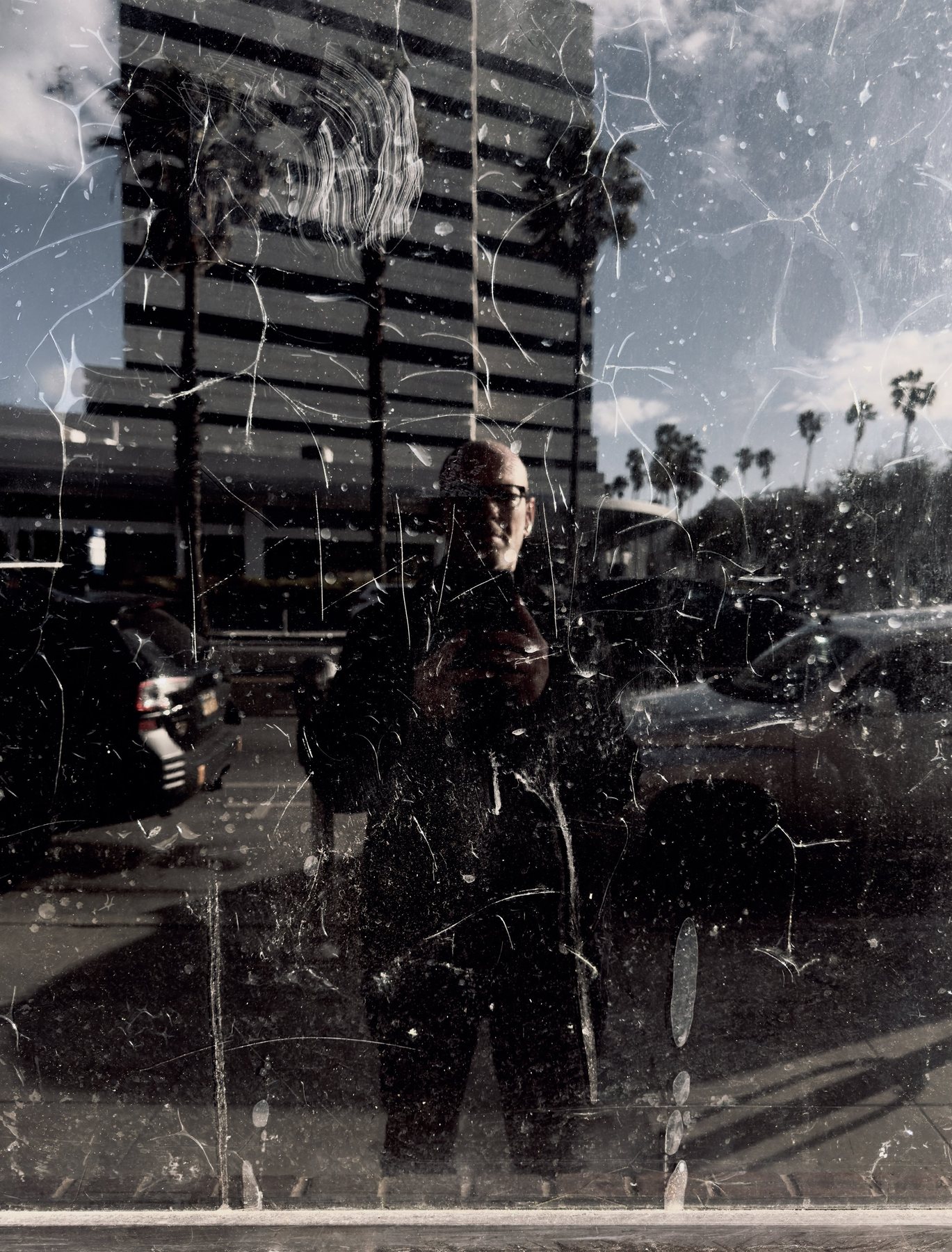 No End In Sight 118: The artist, bespectacled, reflected in scratched, cracked glass with palm trees, a tall building and parked cars behind.