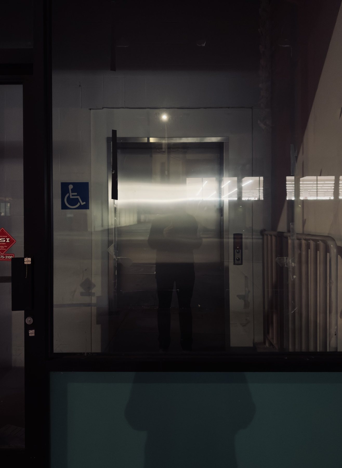 No End In Sight 101: A night window with a disabled access sign and a flash of light, with the artist's dark silhouette standing before an elevator door.