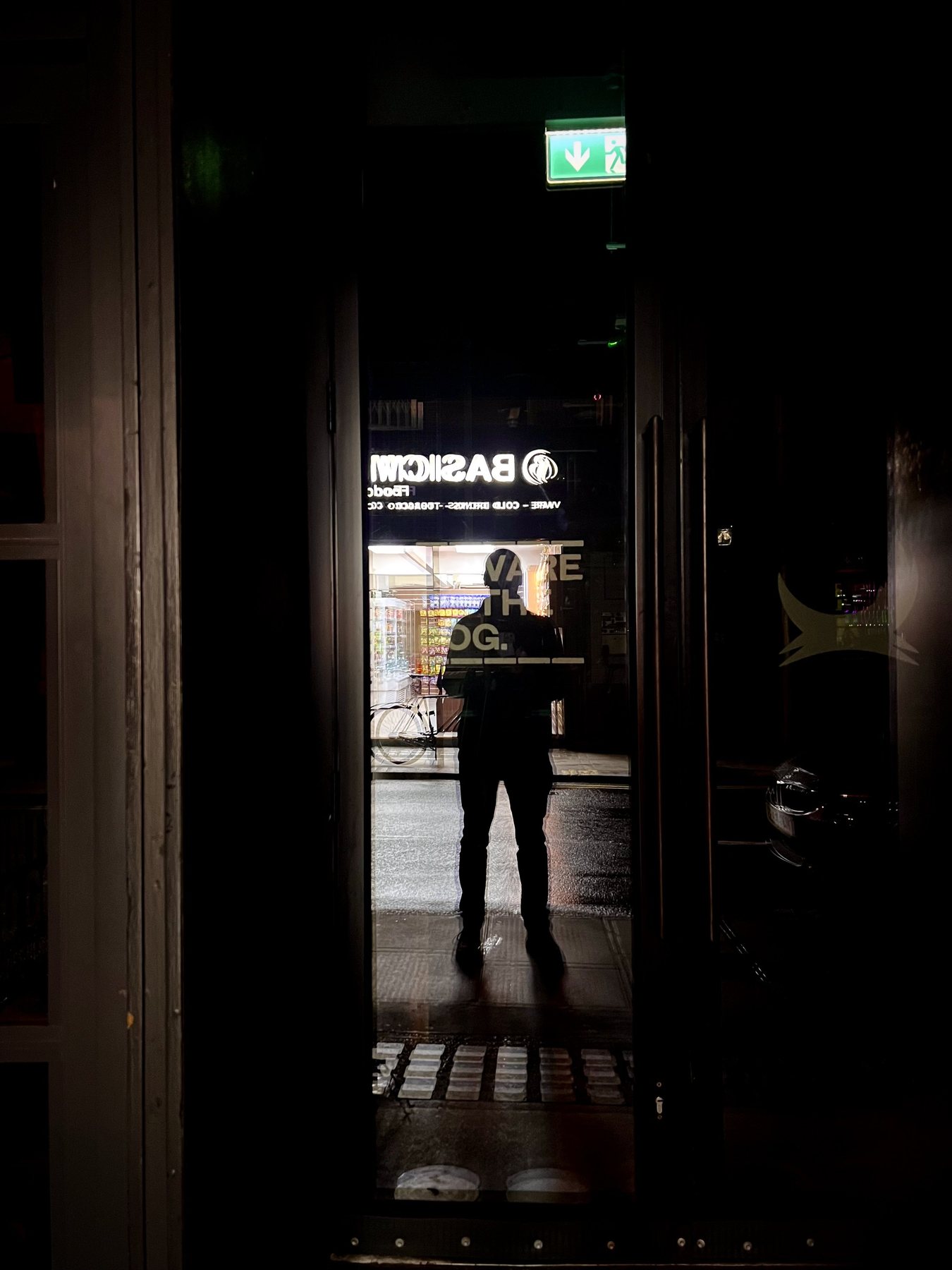 No End In Sight 020: The artist in a dark doorway at night, facing a lit street and a shopfront sign reading BASICM with an exit sign above.