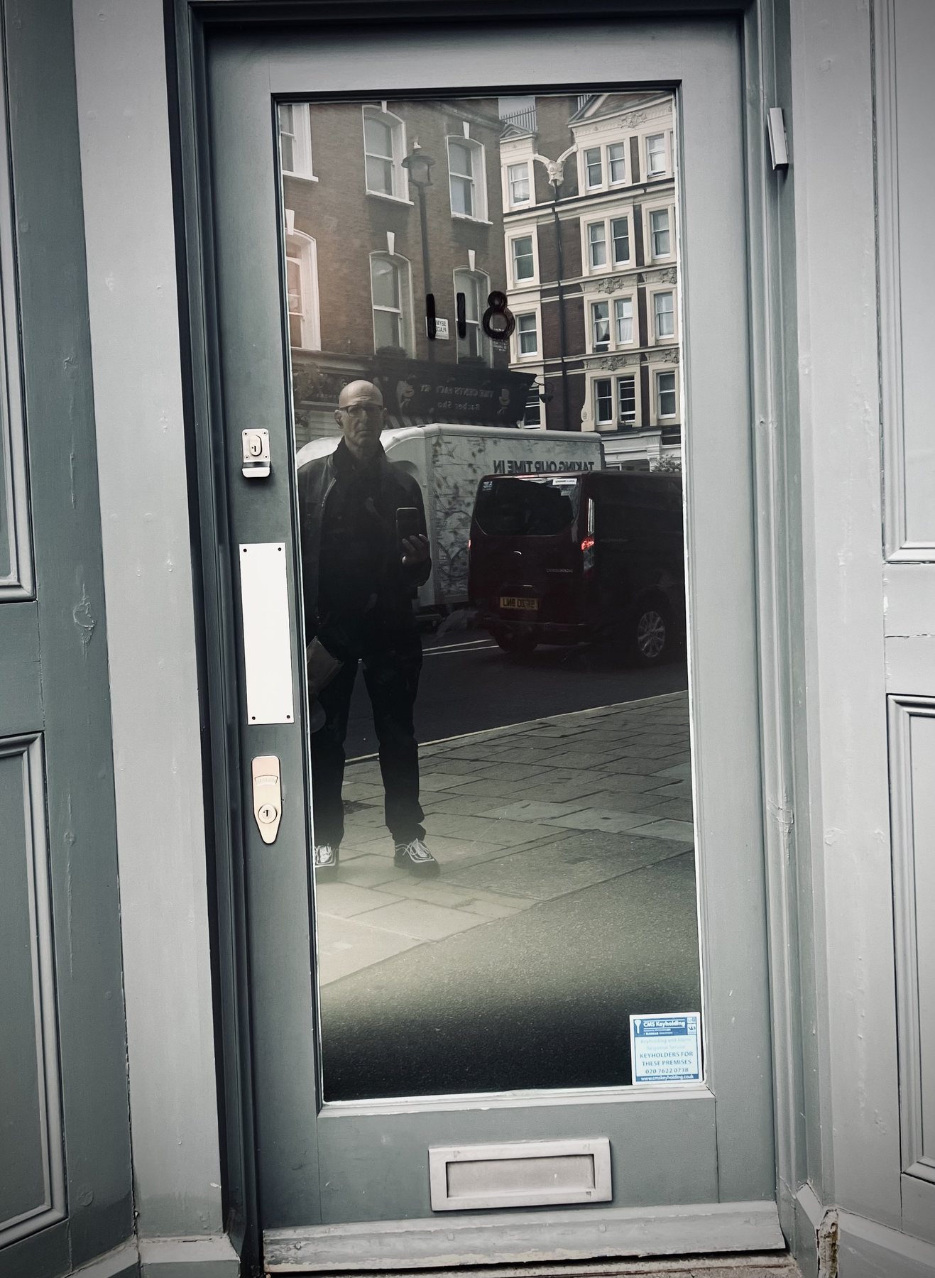 No End In Sight 005: The artist with a phone reflected in the glass of a grey door with letterbox, showing a street, white van and brick buildings.