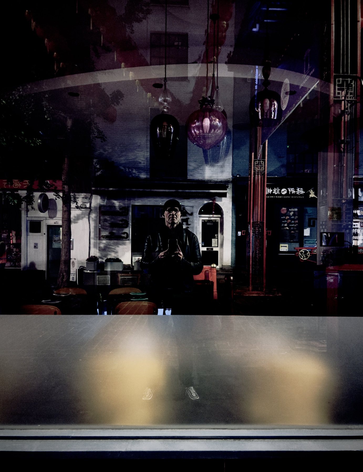 No End In Sight 017: The artist, in a cap and dark jacket, reflected in a restaurant window against red lanterns and Chinese signage across the street.