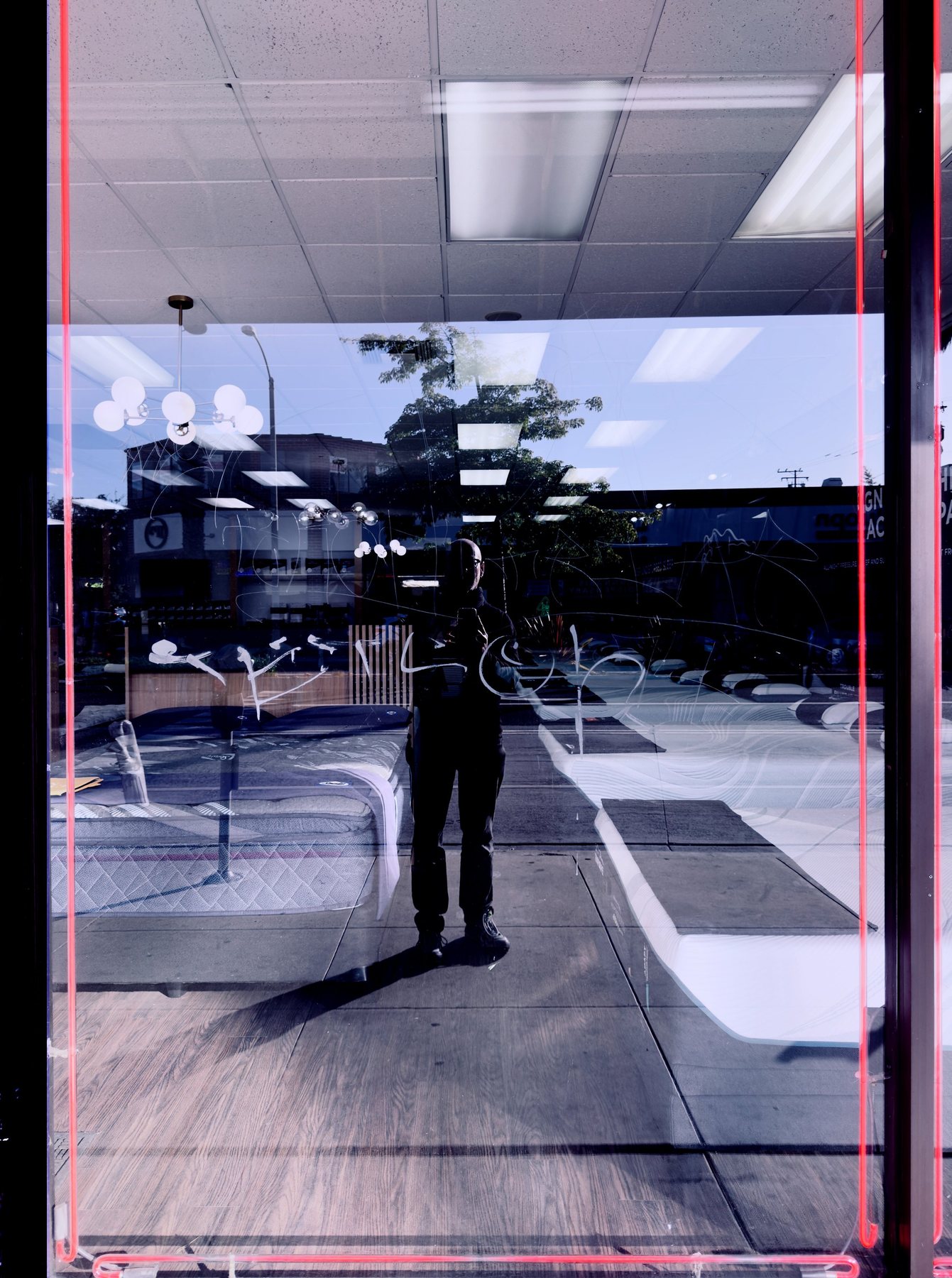 No End In Sight 140: The artist's reflection in a mattress shop window with a pink neon edge, ceiling lights and beds reflected.