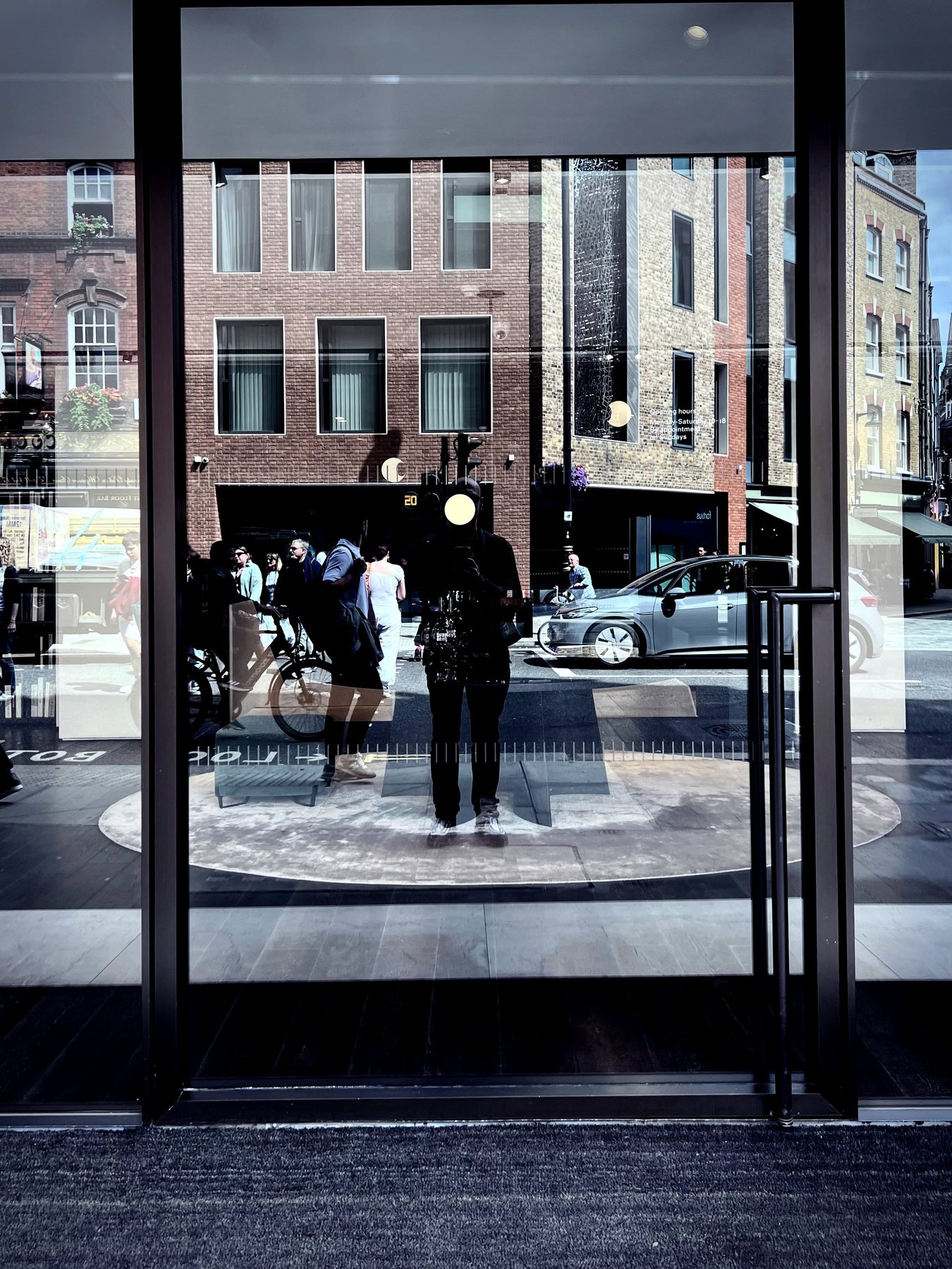 No End In Sight 064: The artist reflected in glass doors with a bicycle, pedestrians and a silver car behind, standing on a circular patterned floor.