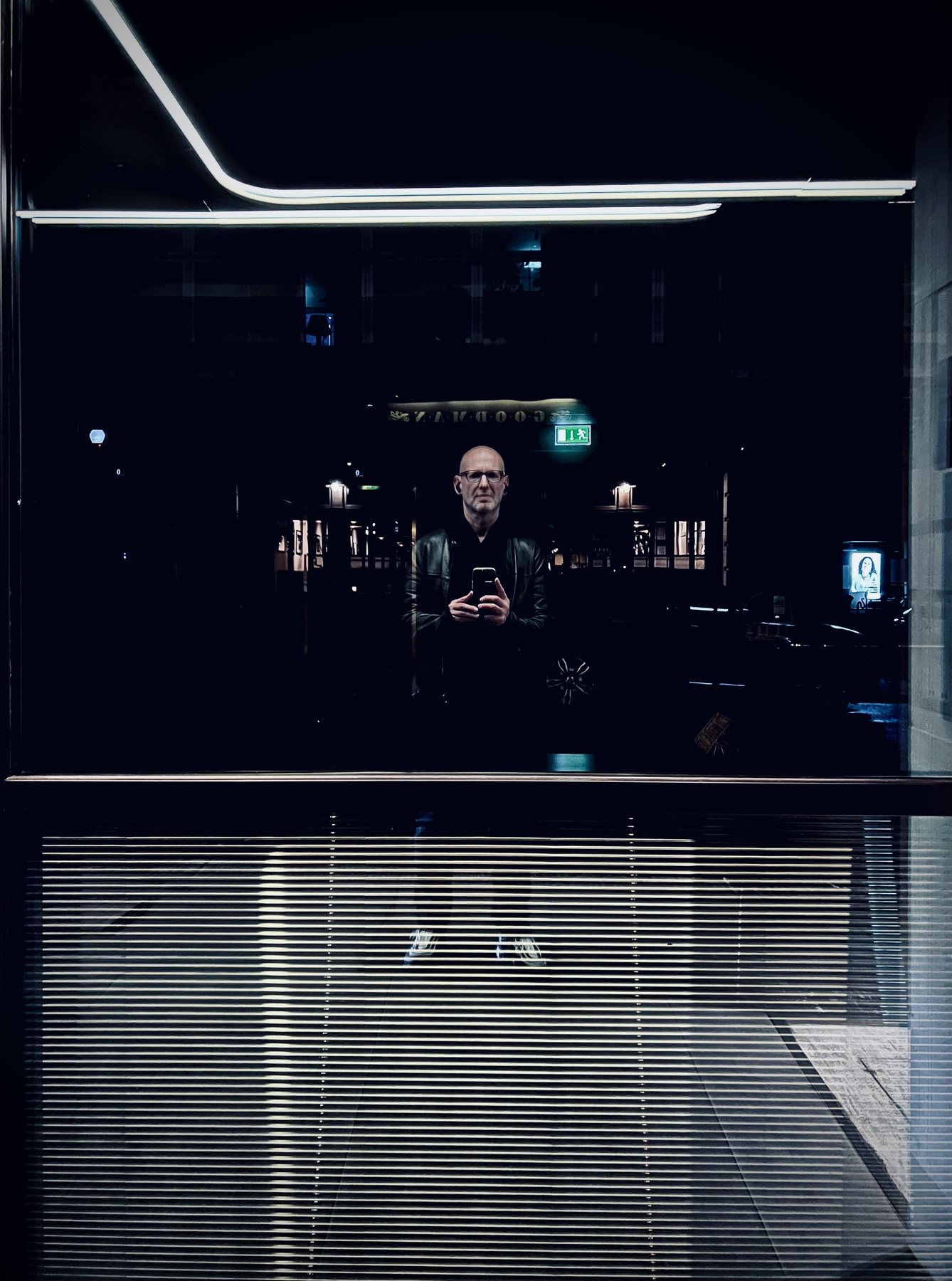 No End In Sight 006: The artist with a phone in a dark window, white light strips above, a green exit sign and a lit street behind.