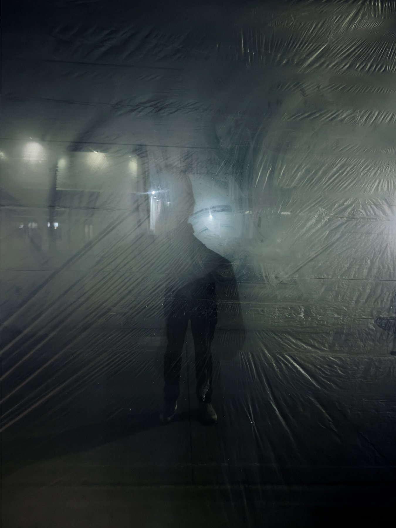 No End In Sight 100: Grey: the artist's dim silhouette behind creased plastic sheeting, with blurred lights glowing in the dark behind.