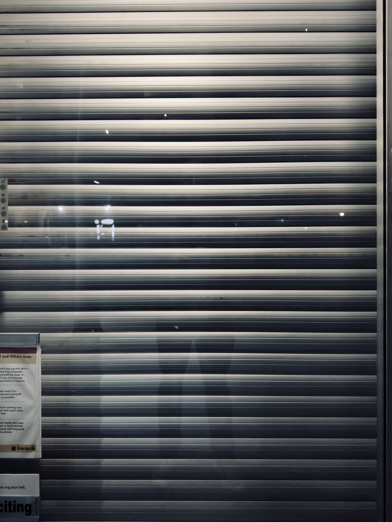 No End In Sight 110: A closed metal roller shutter at night, striped with light, with ghostly legs reflected faintly and a notice posted at lower left.