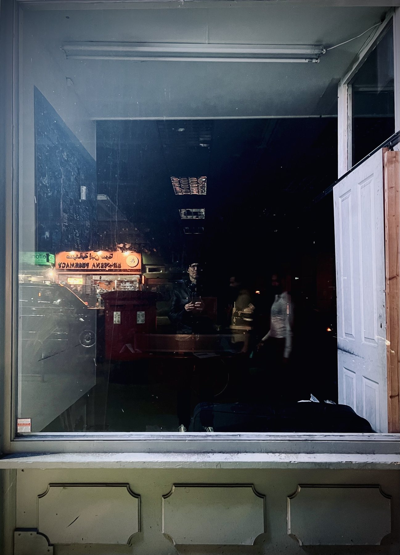 No End In Sight 007: A window showing a shopfront with Arabic signage and a red post box, the artist standing in the dim interior with passersby.