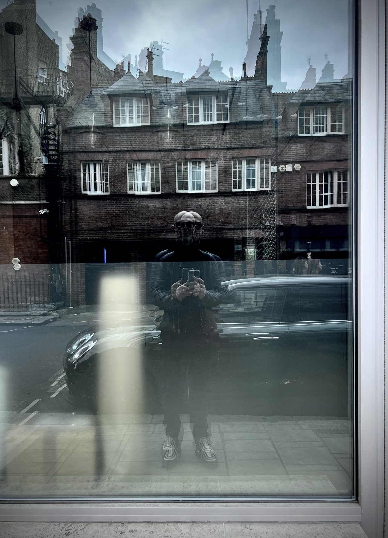 No End In Sight 001: The artist with a phone reflected over a parked dark car in a window with brick terraced houses and layered skyline echoes.