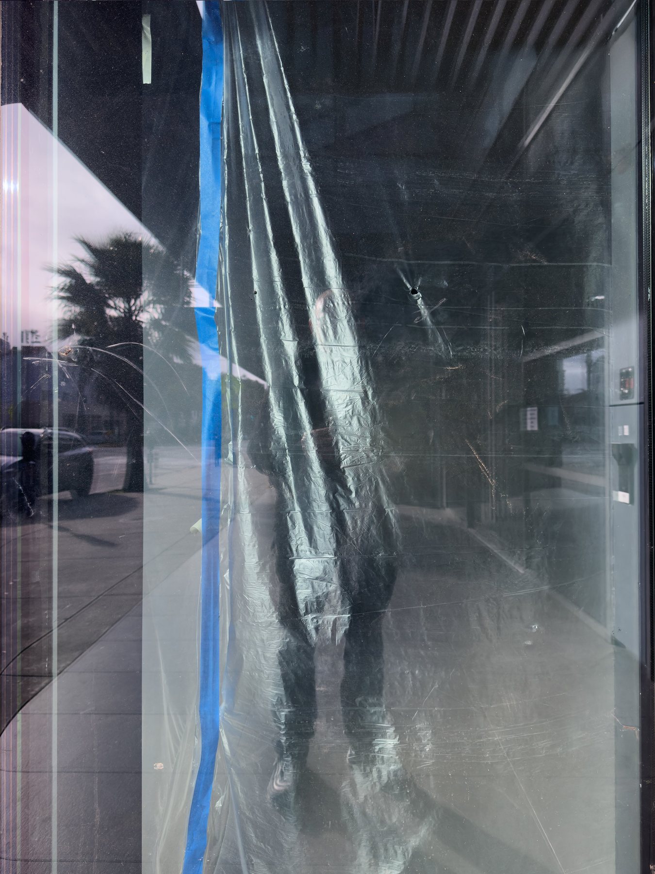 No End In Sight 134: The artist's shape behind creased clear sheeting held by blue tape in a shop window, with a palm tree reflected at left.