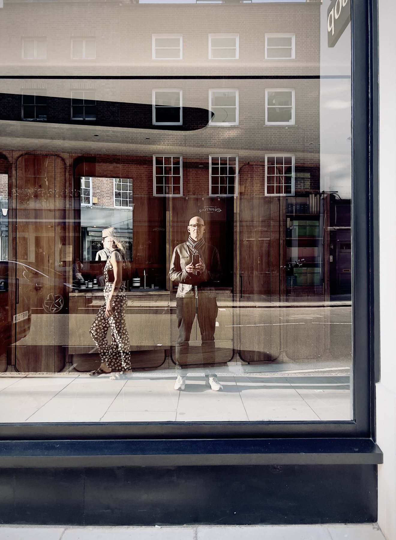 No End In Sight 043: The artist in glasses holding a phone and a woman in patterned trousers walking past, reflected in a shop window with a wooden interior.