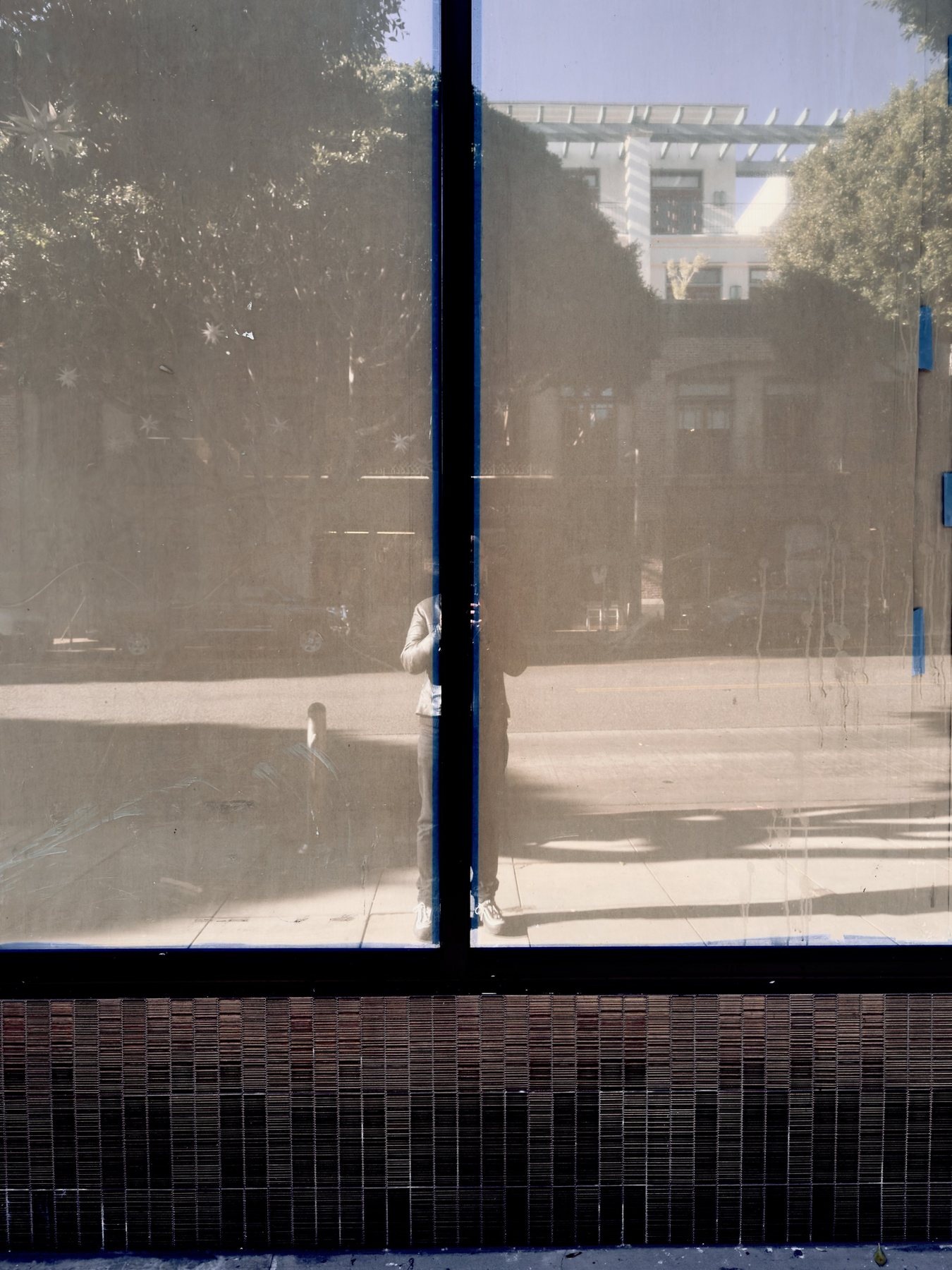No End In Sight 087: The artist reflected in sepia-toned glass with blue edging, trees, a white building and a tiled base beneath the window.