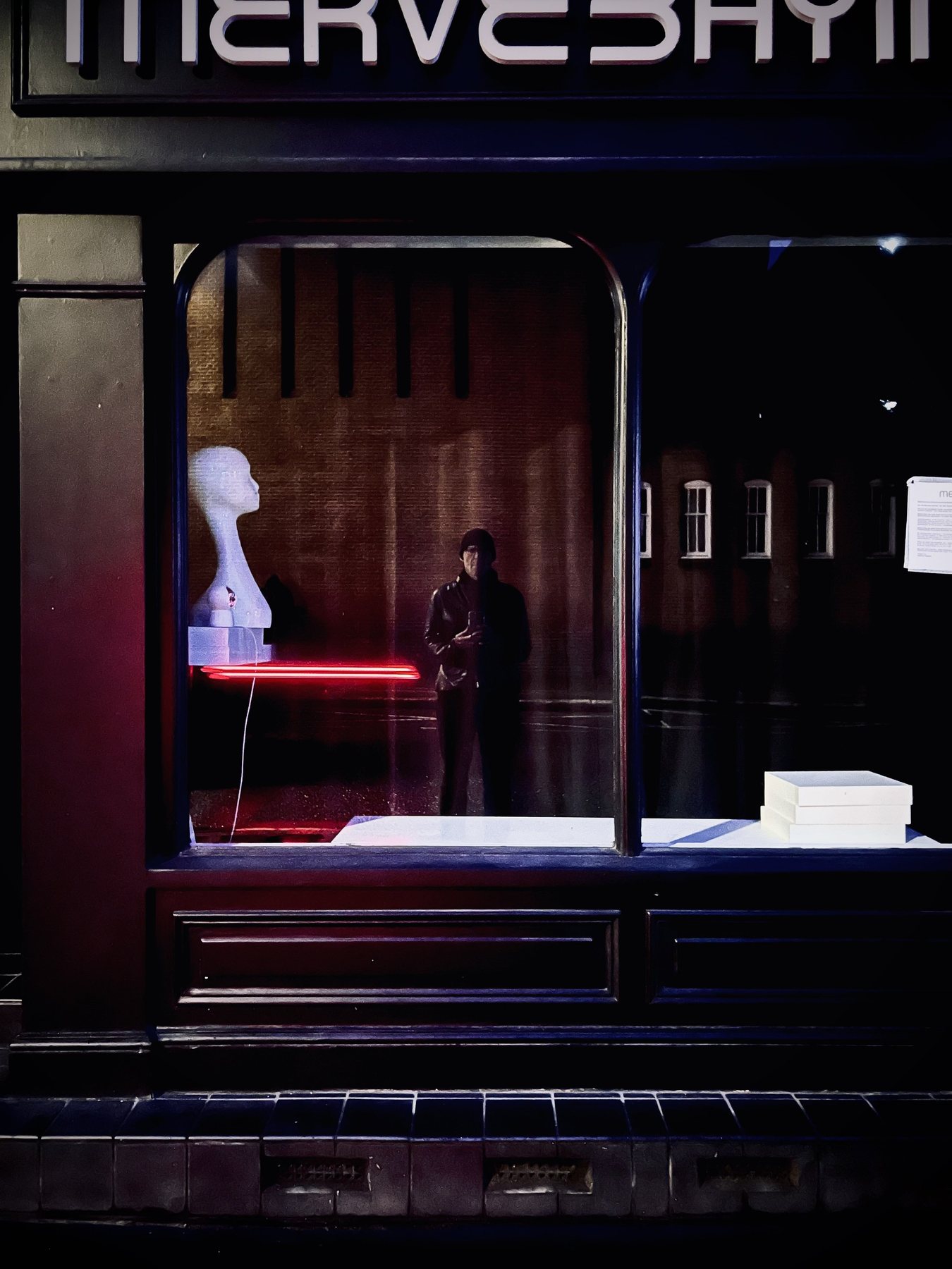 No End In Sight 014: A dark shopfront with a white mannequin head, a red neon tube and the artist in a cap standing in the window.