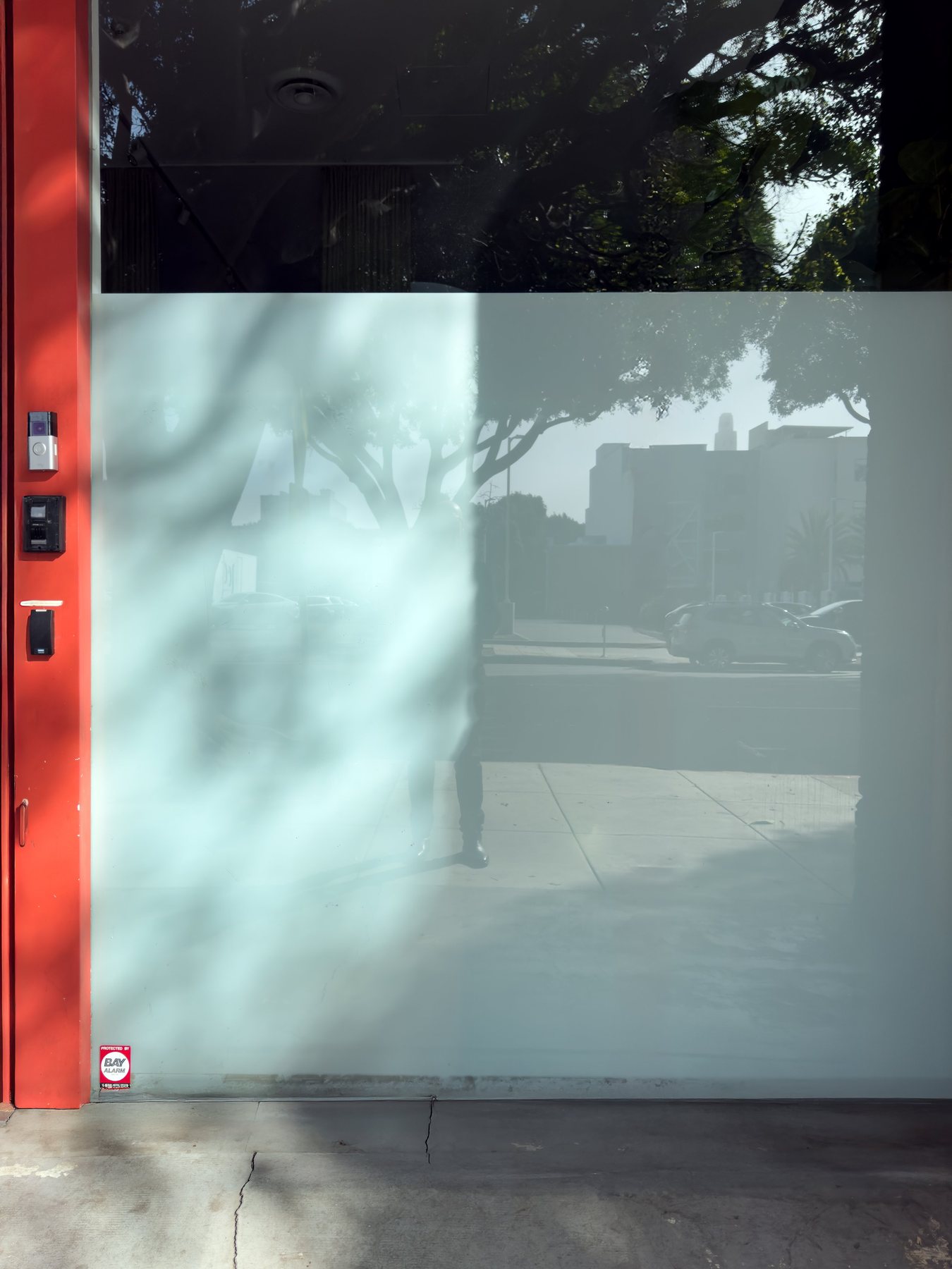 No End In Sight 102: The artist's pale ghostly reflection in a frosted glass panel, with a red door frame, trees and a road with cars.