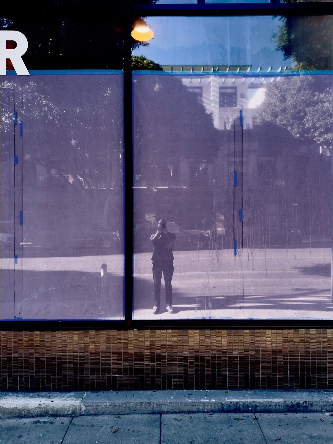 No End In Sight 093: The artist reflected in purple-tinted window film with blue tape marks, a letter R and a sunlit street with trees.