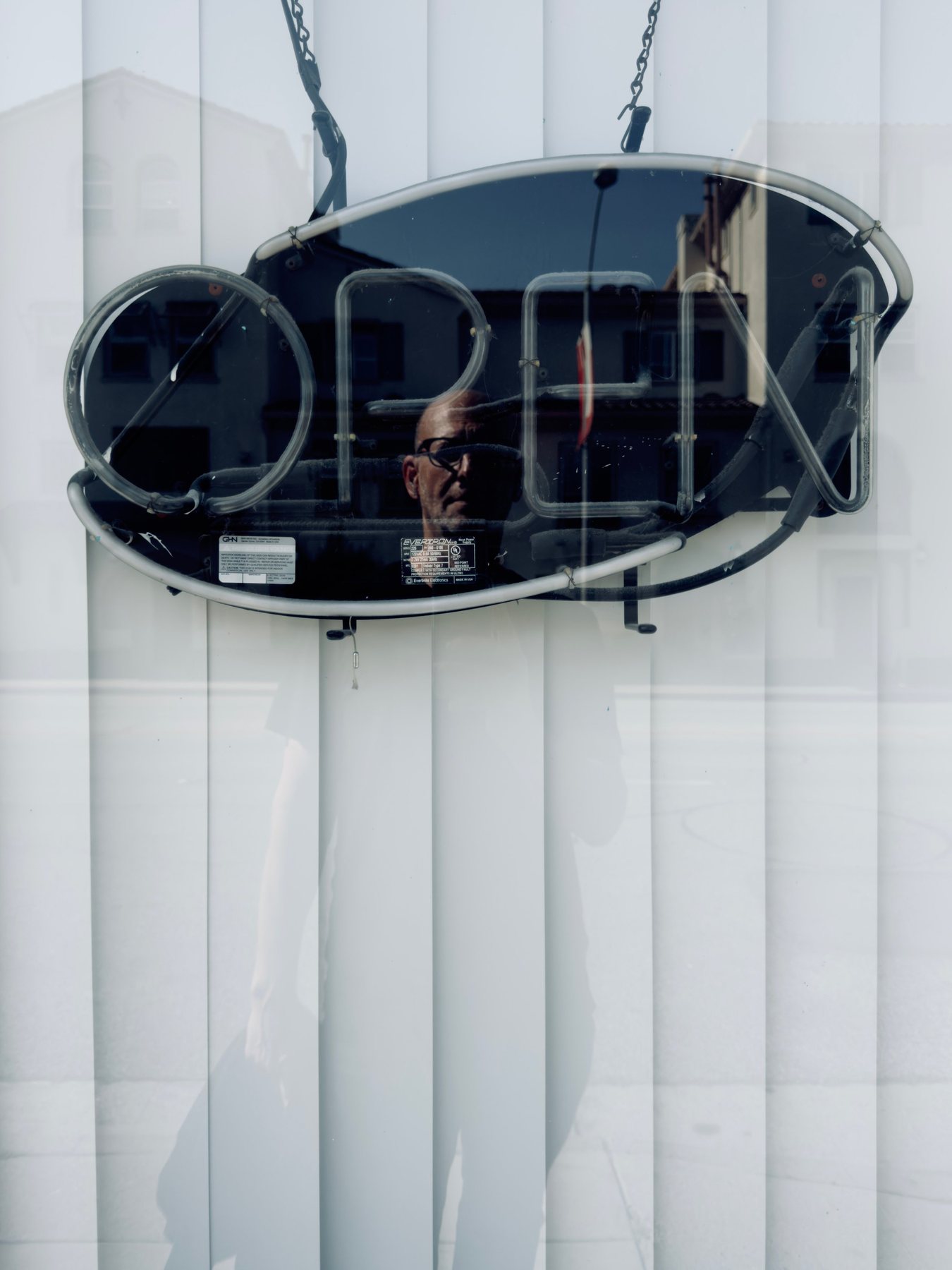 No End In Sight 085: The artist's face reflected inside the neon OPEN sign, hanging in a window with white vertical blinds and a faint standing silhouette.