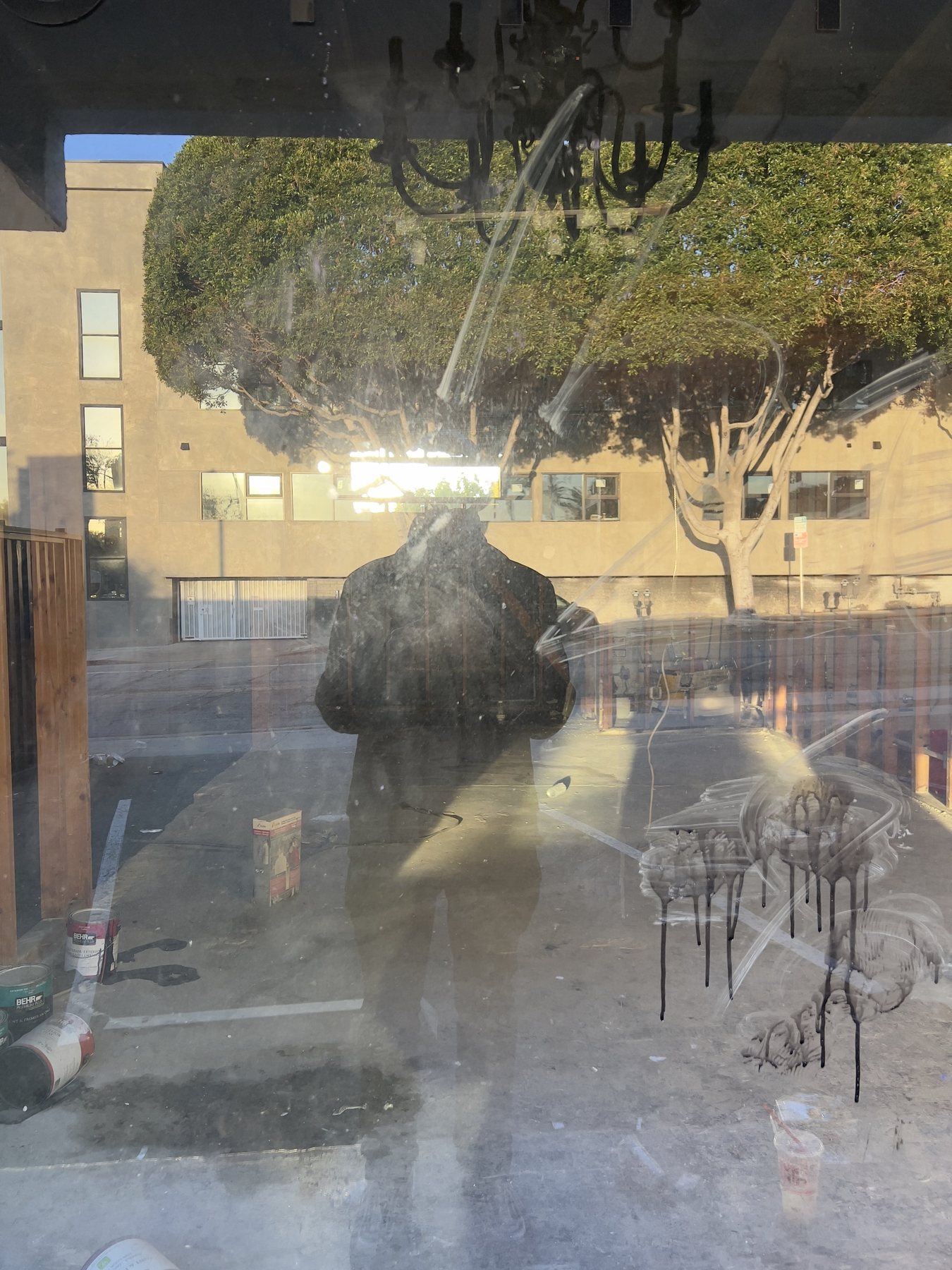 No End In Sight 076: A dirty window reflecting a leafy tree and the artist's dark figure, with paint cans and white drips on the glass, a chandelier above.