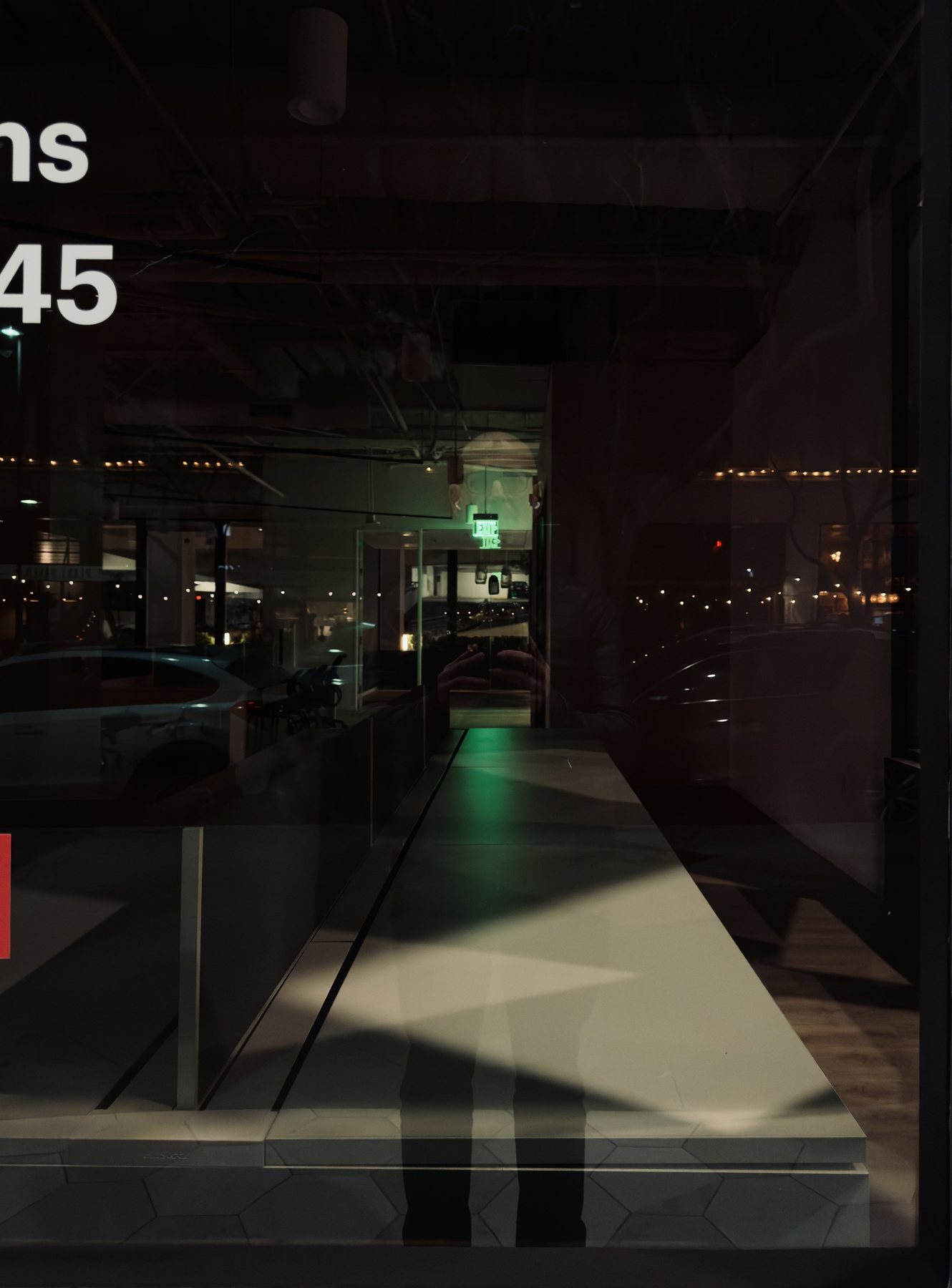 No End In Sight 109: Night shopfront window with a green-lit counter and string lights, a parked car reflected, partial white lettering and a dark figure's legs below.