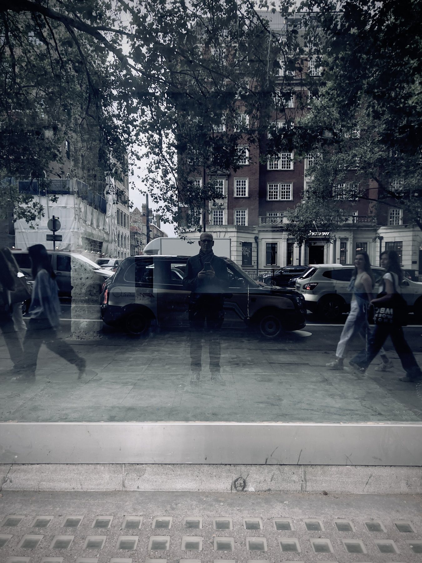 No End In Sight 041: A monochrome window reflecting a black taxi, trees, a brick building, pedestrians and the artist above a paved pavement.