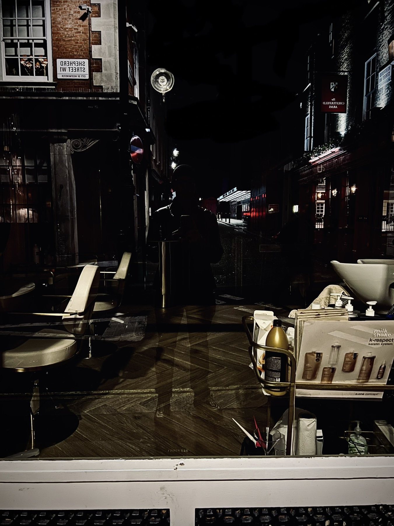 No End In Sight 012: A night window onto a hairdresser's with white chairs and a basin, the artist's silhouette against a street sign reading Shepherd Street.