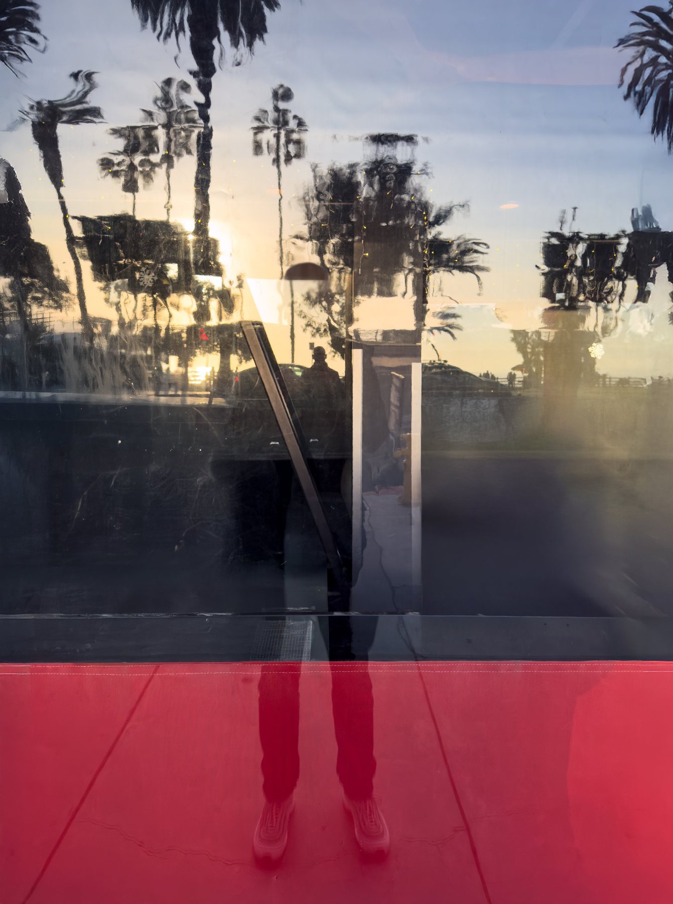 No End In Sight 095: The artist's legs and shoes below a red band, with palm trees and a sunset reflected in the glass above a pavement.