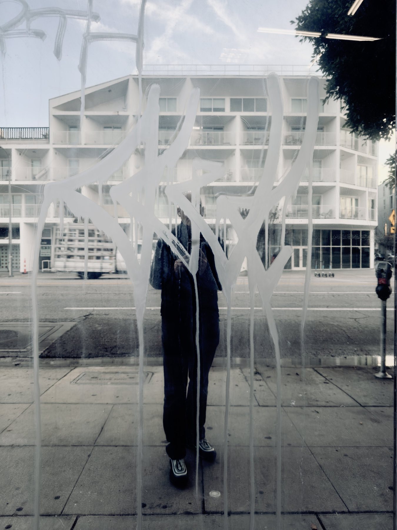 No End In Sight 121: The artist's reflection behind a white spray-painted star tag on glass, with a pale modern apartment building across the street.