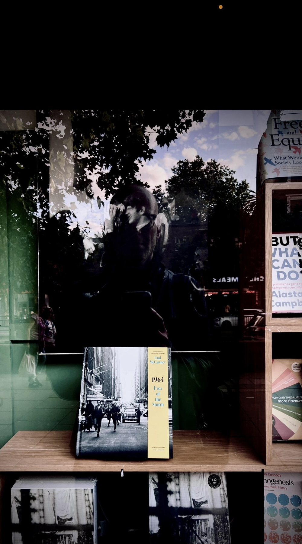 No End In Sight 094: The artist's reflection in a bookshop window above a displayed book with a street photograph on its cover, with trees and a street in the glass.