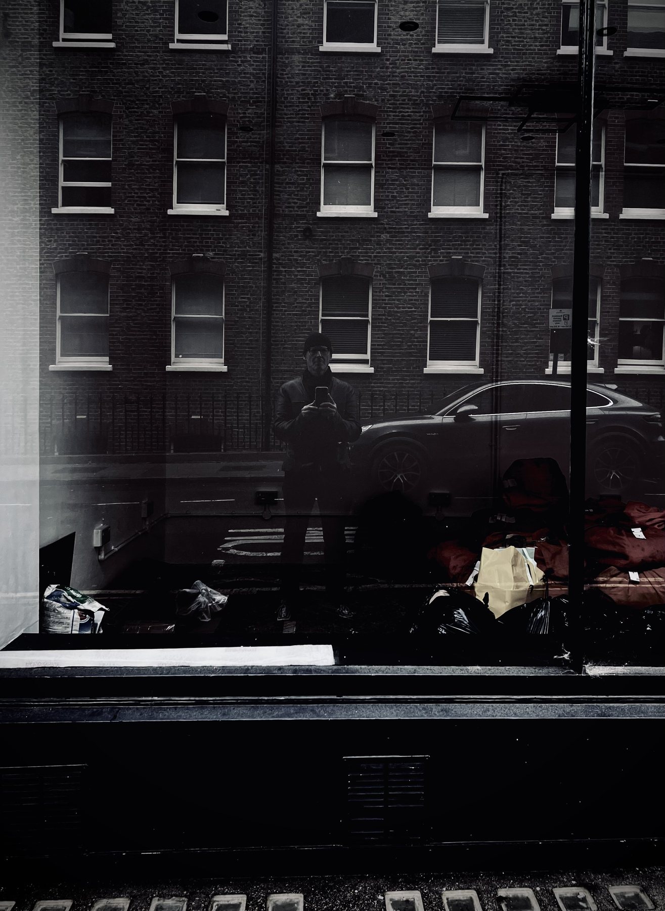 No End In Sight 011: The artist reflected in a dark window with a brick terrace, a parked car and bags of rubbish on the pavement.