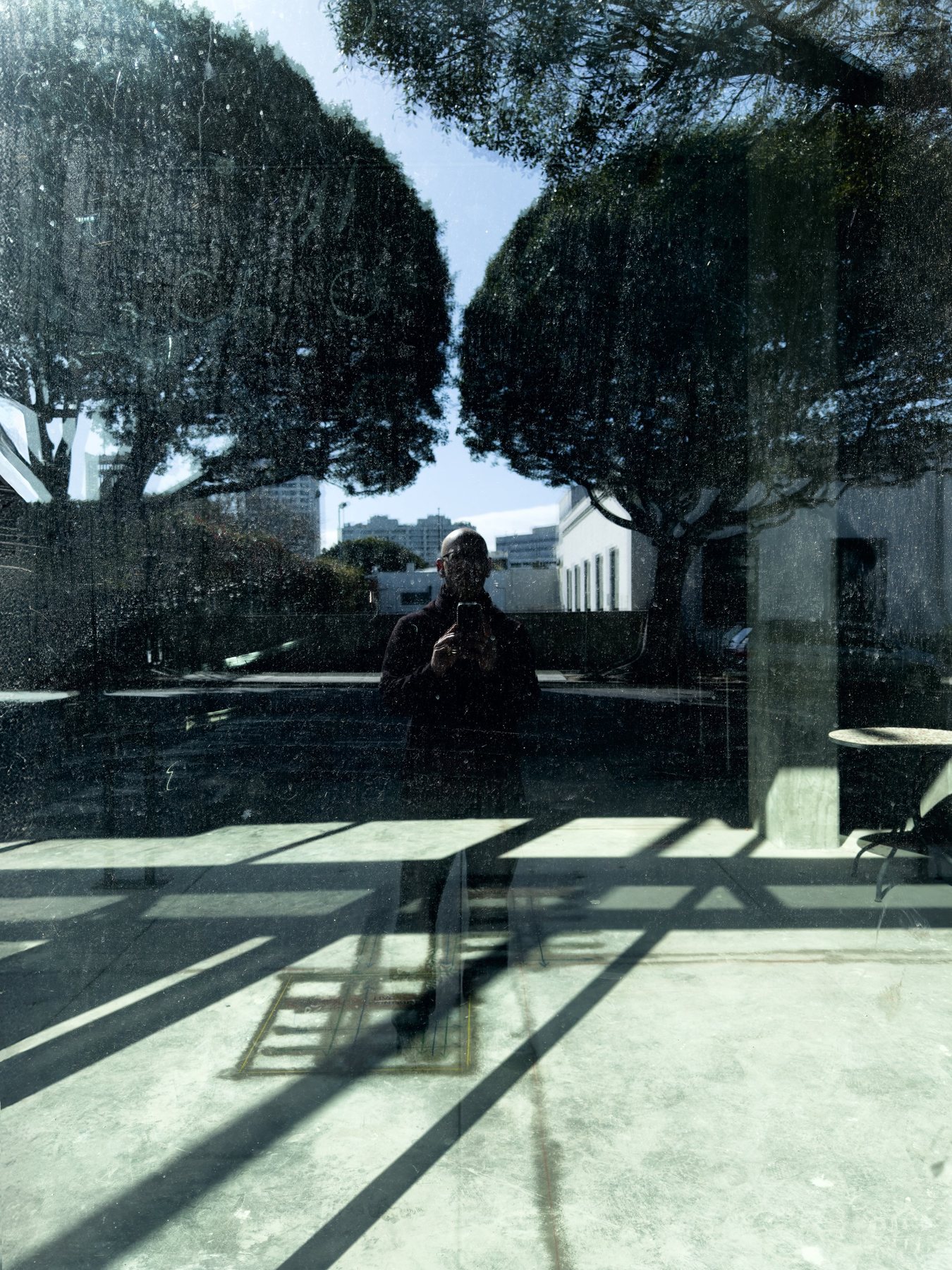 No End In Sight 136: The artist holding a phone, reflected in dark glass with two rounded trees, sunlit paving and long diagonal shadows.