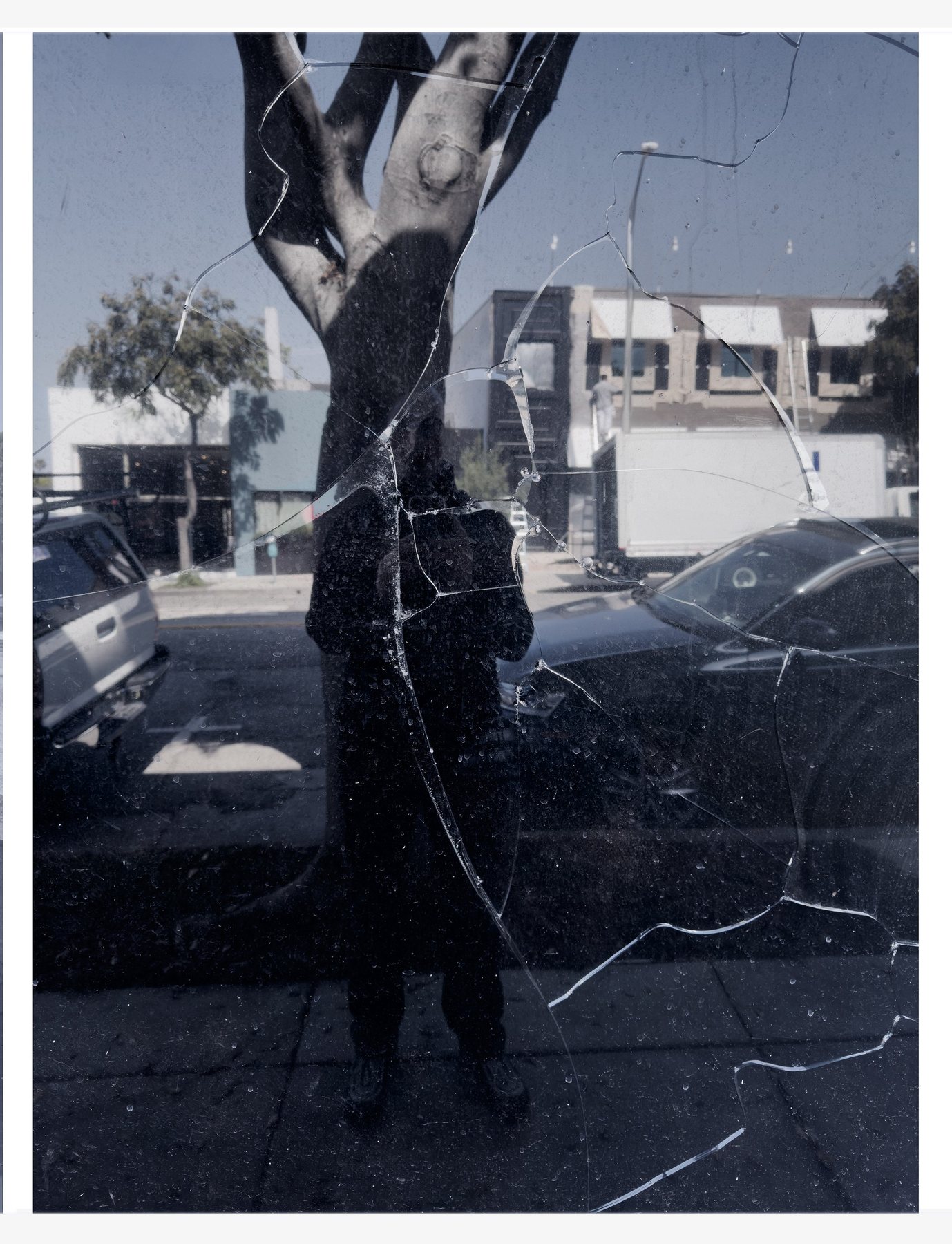 No End In Sight 144: The artist's reflection behind a cracked pane of glass, with a tree trunk, parked cars and a white truck on the street.