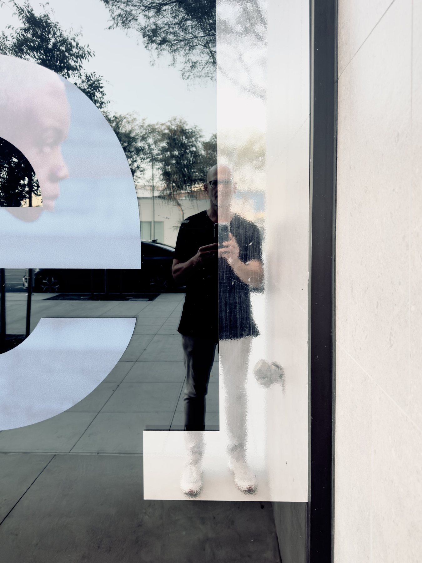 No End In Sight 083: The artist in a dark t-shirt and white trousers reflected in a window with large white letterforms and a portrait fragment.