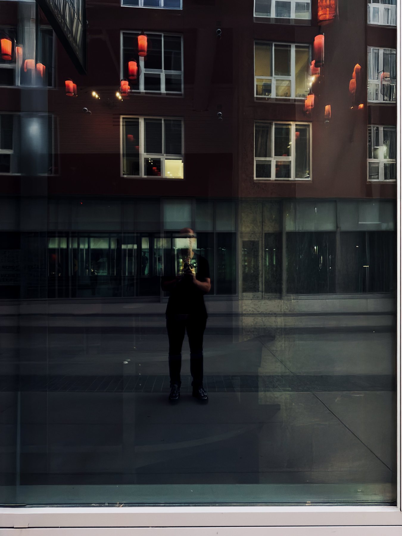 No End In Sight 130: The artist holding a phone, reflected in a window over a brick building hung with red lanterns.