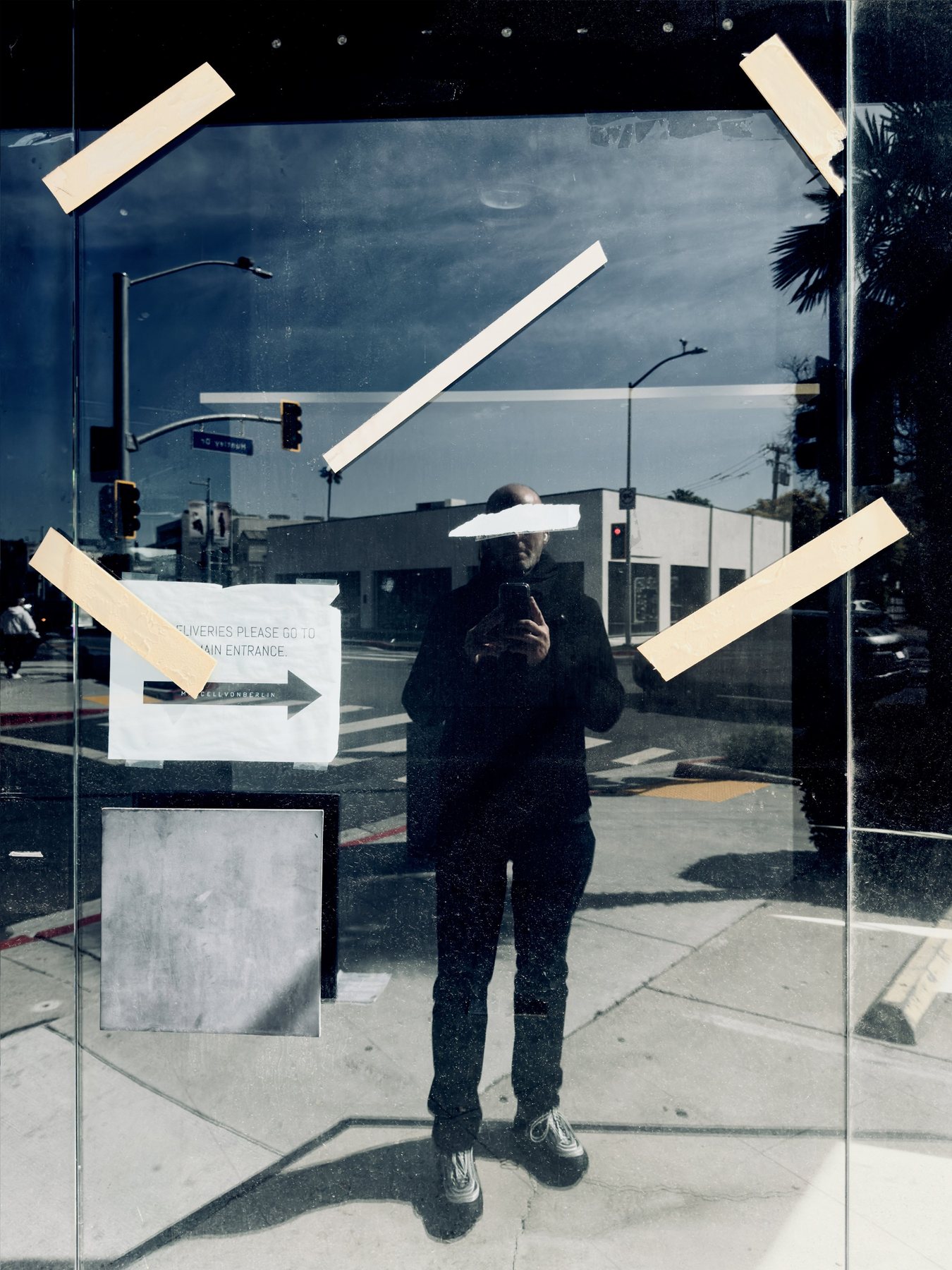 No End In Sight 115: The artist reflected in a taped glass door, with strips of tape across the glass, traffic lights, a street corner and a directional sign.