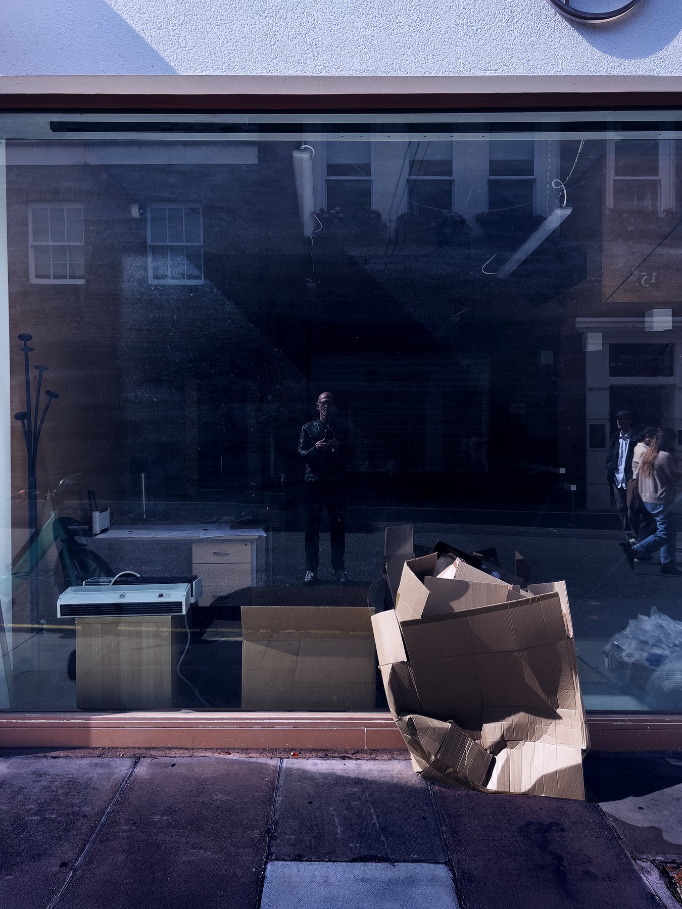 No End In Sight 052: The artist's faint reflection in an empty shop window with a desk, a printer and flattened cardboard boxes on the pavement.