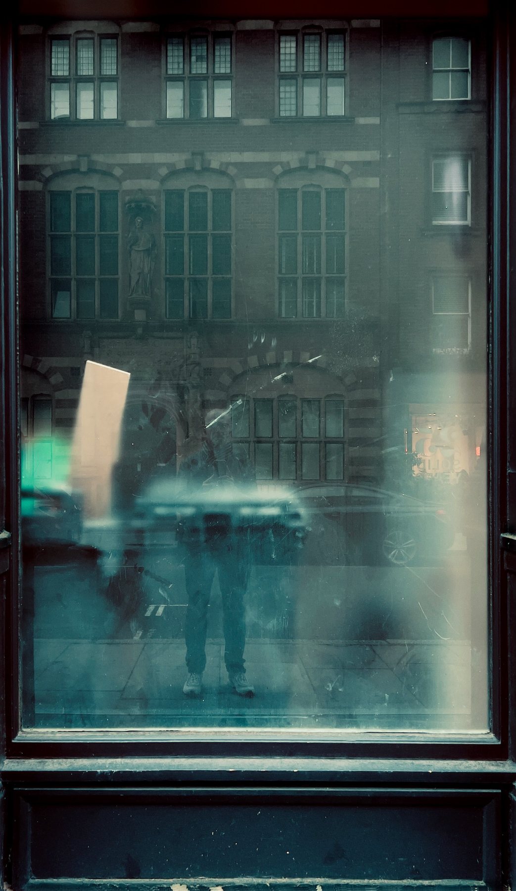 No End In Sight 048: A teal-toned window with a ghostly lower-body reflection and a brick building with tall windows, the glass streaked and dusty.
