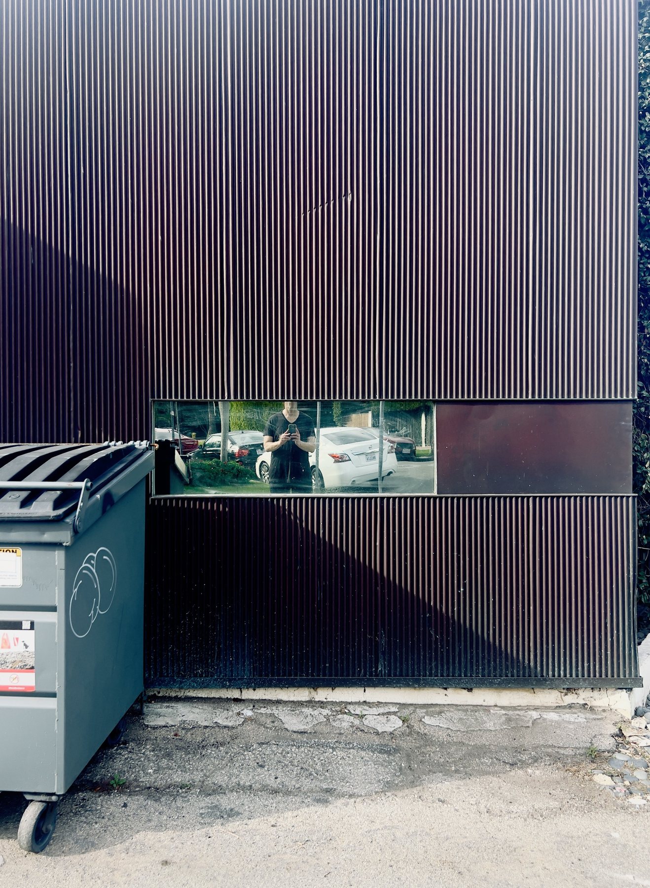 No End In Sight 065: The artist with a phone reflected in a narrow window slot in a corrugated dark wall, beside a green dumpster and parked white cars.