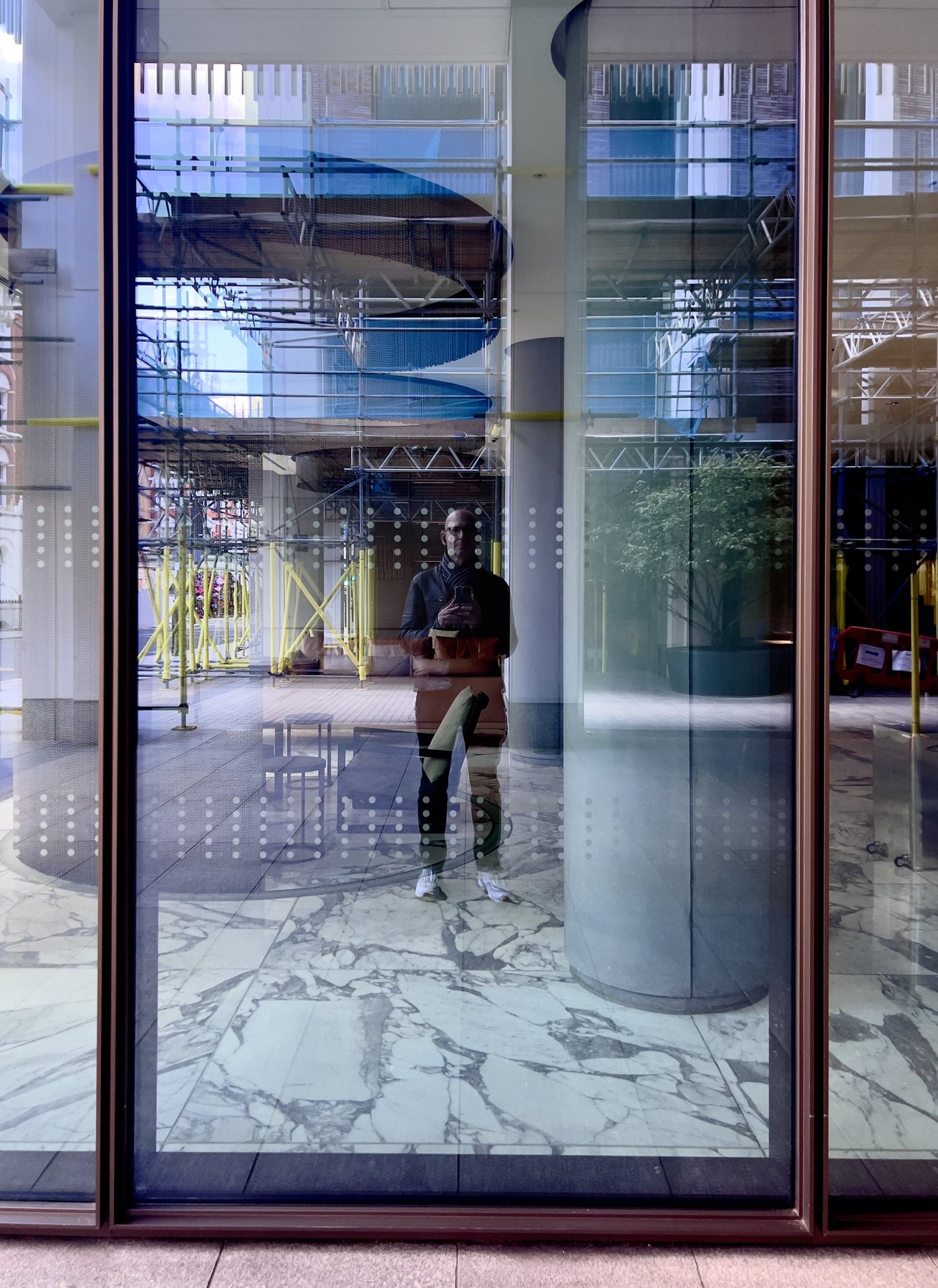 No End In Sight 045: The artist reflected in a tall glass facade with scaffolding, a curved column and a marble floor, dotted frosting across the glass.