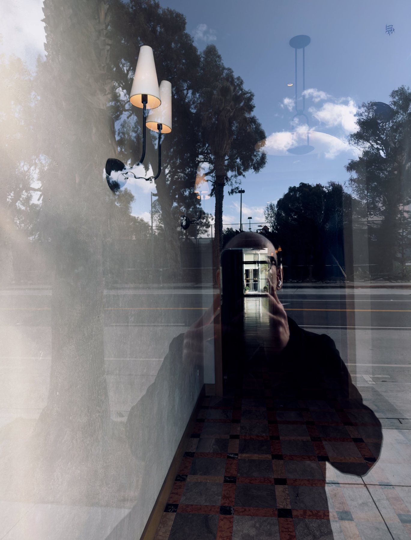 No End In Sight 119: The artist's reflection in a window overlaid with a view of trees, a wall lamp with two shades and a tiled floor.