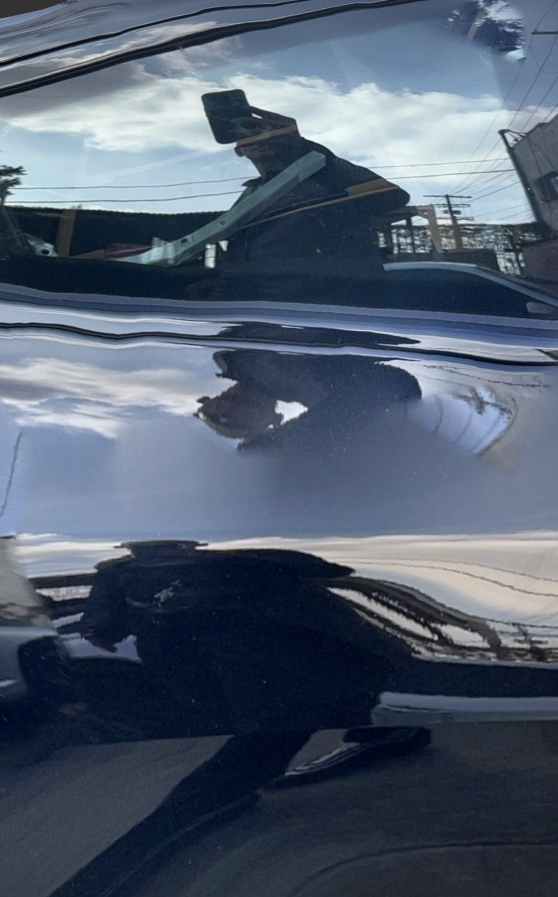 No End In Sight 073: The artist's reflection warped across the glossy black surface of a car, with blue sky, clouds, power lines and the artist's phone.