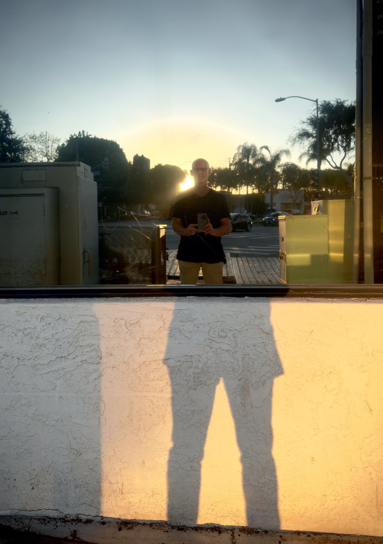 No End In Sight 084: The artist photographing his reflection in a window at sunset, his long shadow falling across a white stucco wall below.