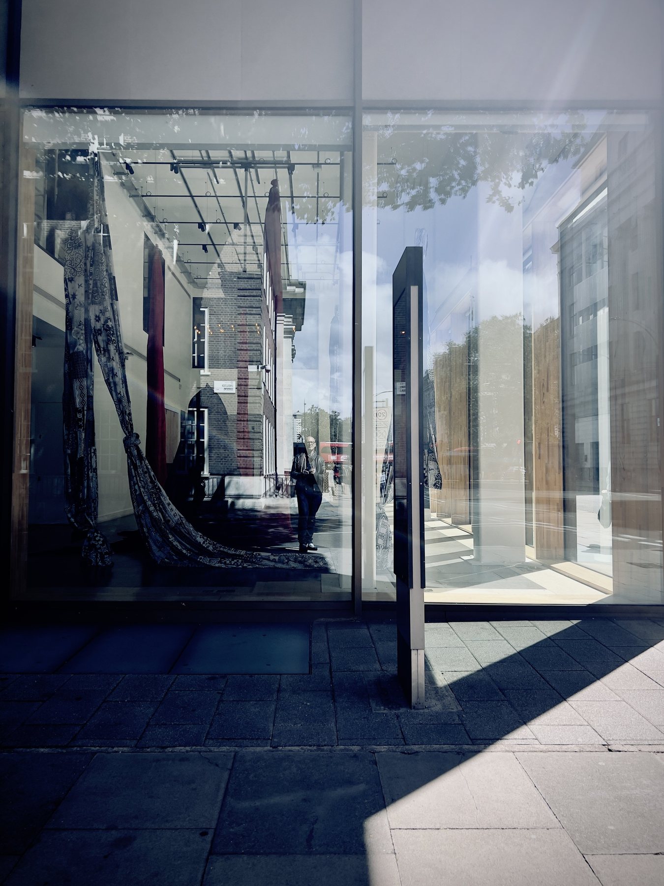 No End In Sight 049: A sunlit shop window reflecting a street, with the artist's dark figure and a rippled hanging fabric inside the building.