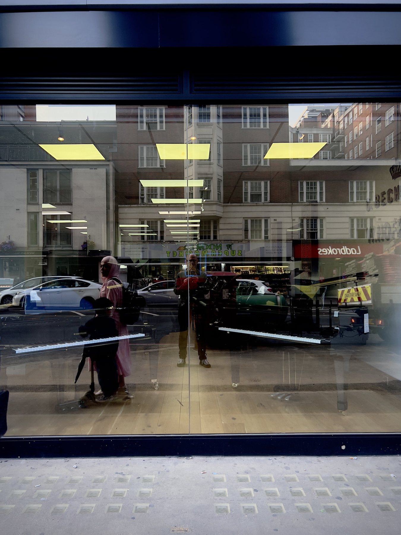No End In Sight 026: The artist and a passerby in a pink coat reflected in a daytime London shopfront window, with a Ladbrokes sign and keyboards inside.