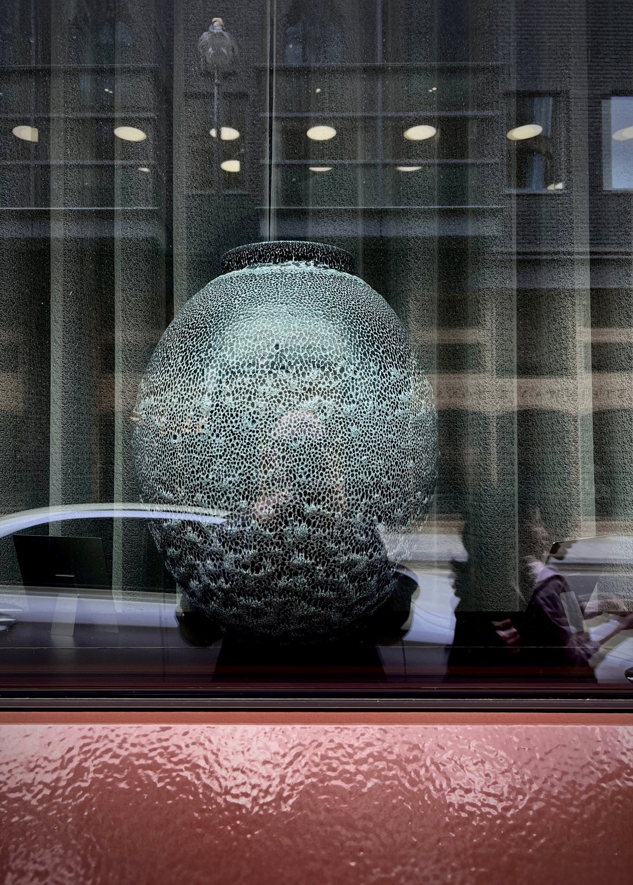 No End In Sight 013: A large ceramic-like globe with a cracked, speckled surface, seen through a window with a pink-red sill and passers-by reflected.