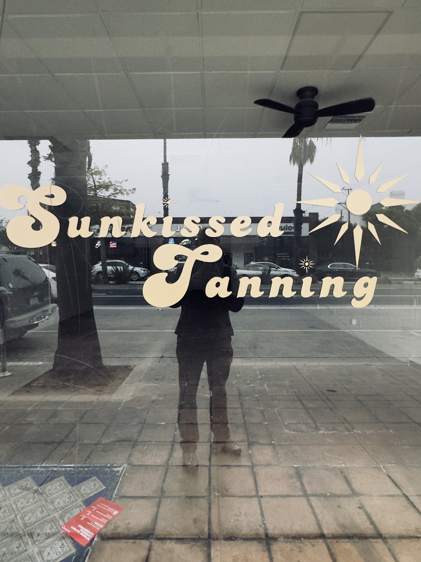 No End In Sight 124: The artist's reflection behind cream vintage lettering reading Sunkissed Tanning, with a ceiling fan, palm tree and street behind.