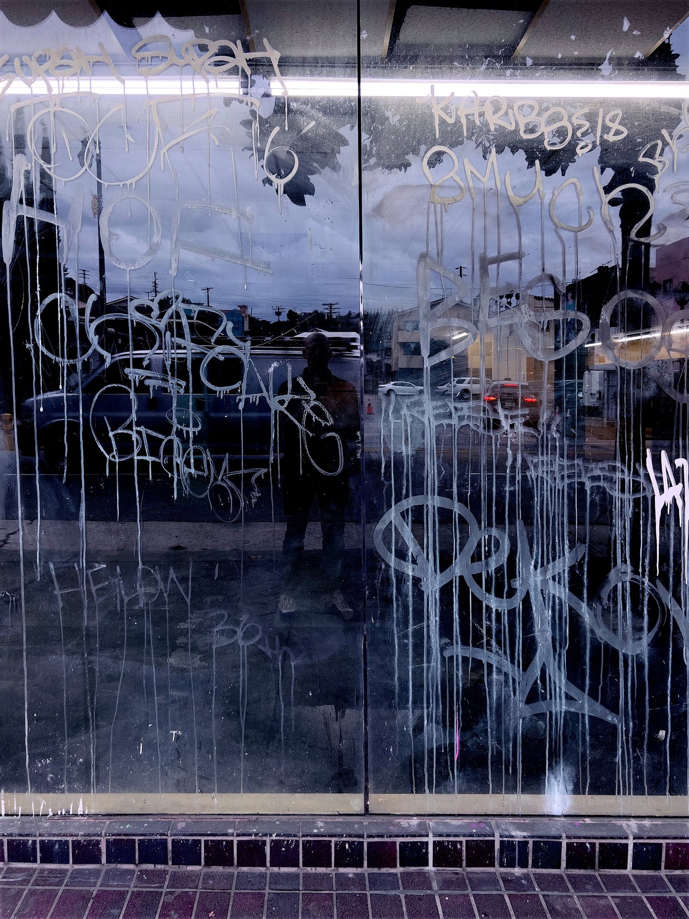 No End In Sight 059: The artist's dim reflection in a shopfront covered with white graffiti and drips, with a purple-tiled sill below.