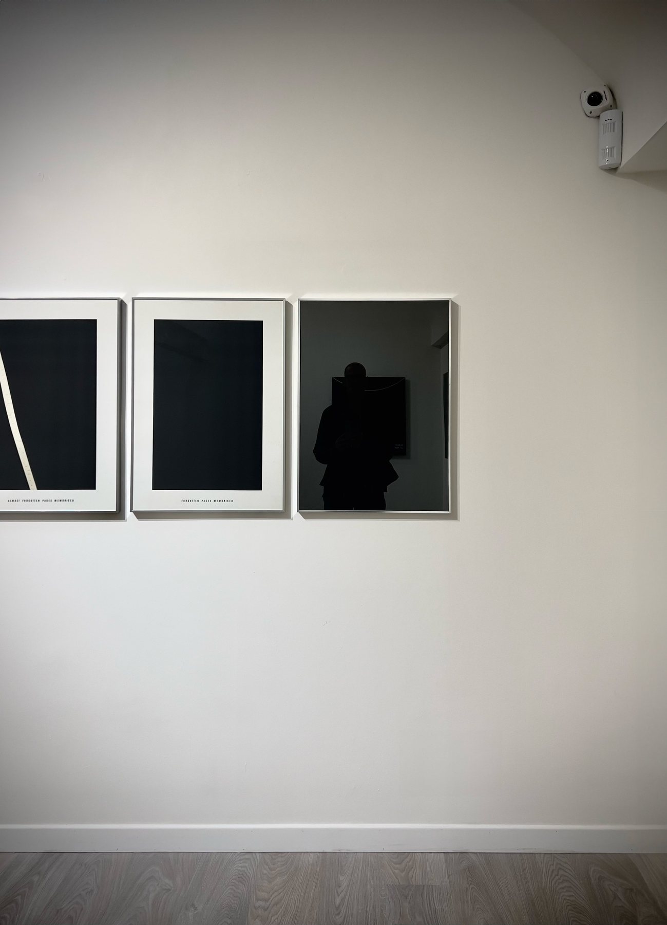 No End In Sight 056: The artist's reflection in a black mirror panel on a white gallery wall, beside two framed dark prints.